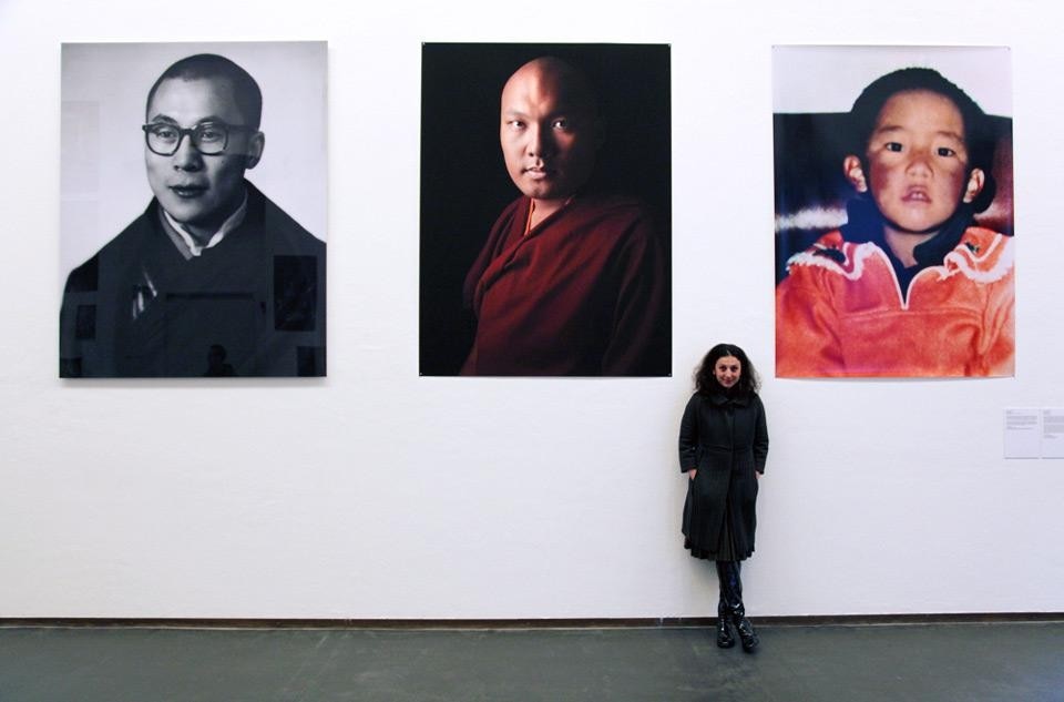 Paola Pivi, <em>Tulkus 1880 to 2018</em>, 2012. Portrait of the artist. In the background, left, the 14th Dalai Lama at the age of 22, in a 1957 portrait by Kinsey Brothers, Delhi. Courtesy of the Newark Museum. Centre, the 17th Karmapa in a 2012 photo by David Zimmerman. Right, the eleventh Panchen Lama, who was made to disappear by the Chinese government only three days after he was recognized by His Holiness the Dalai Lama in 1995