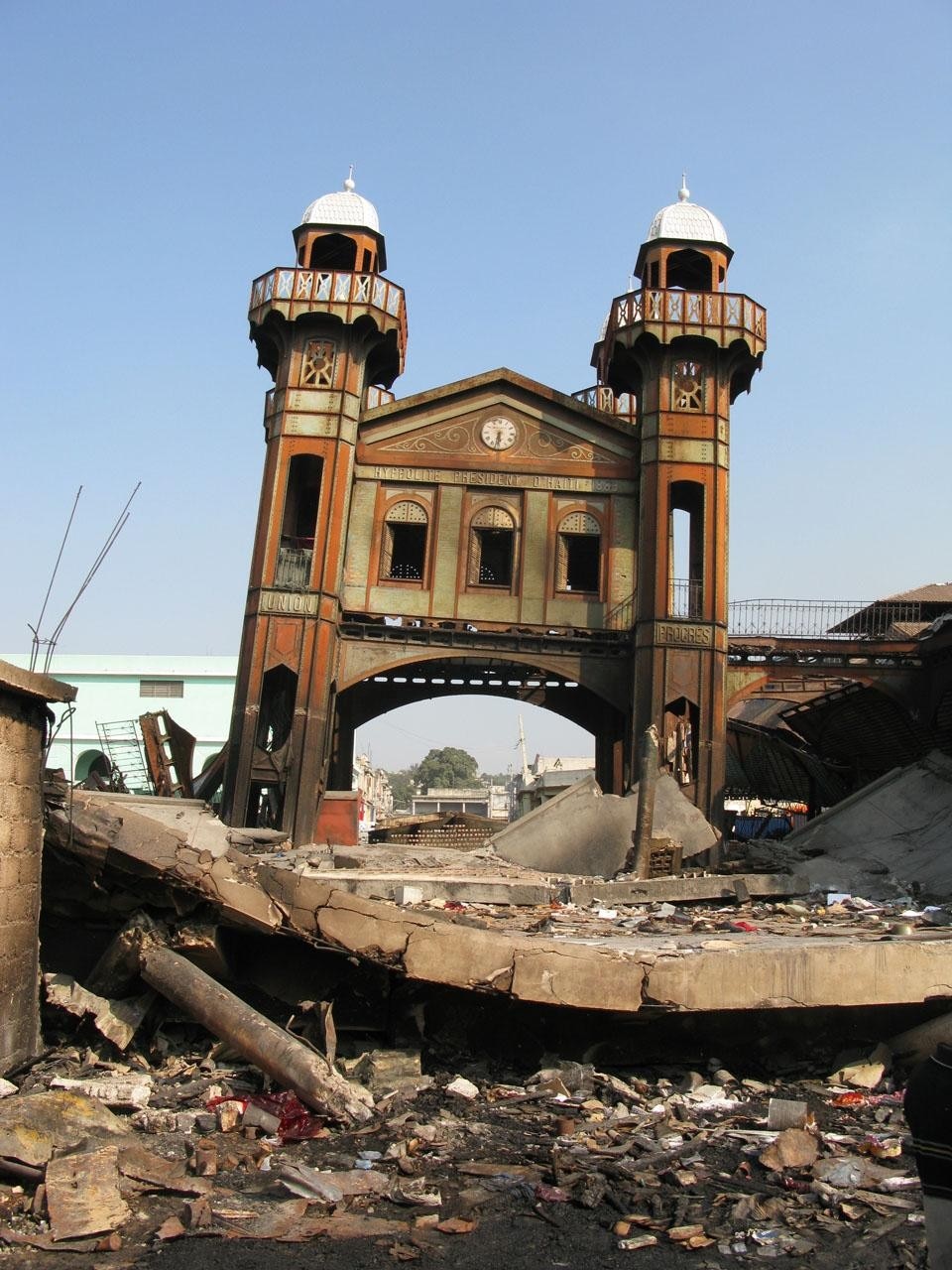 Shortly after the earthquake on 12 Jan 2010, amid the rubble, the two red and green wrought iron towers, slightly skewed, burnt and broken, are still upright and connected to two covered halls.