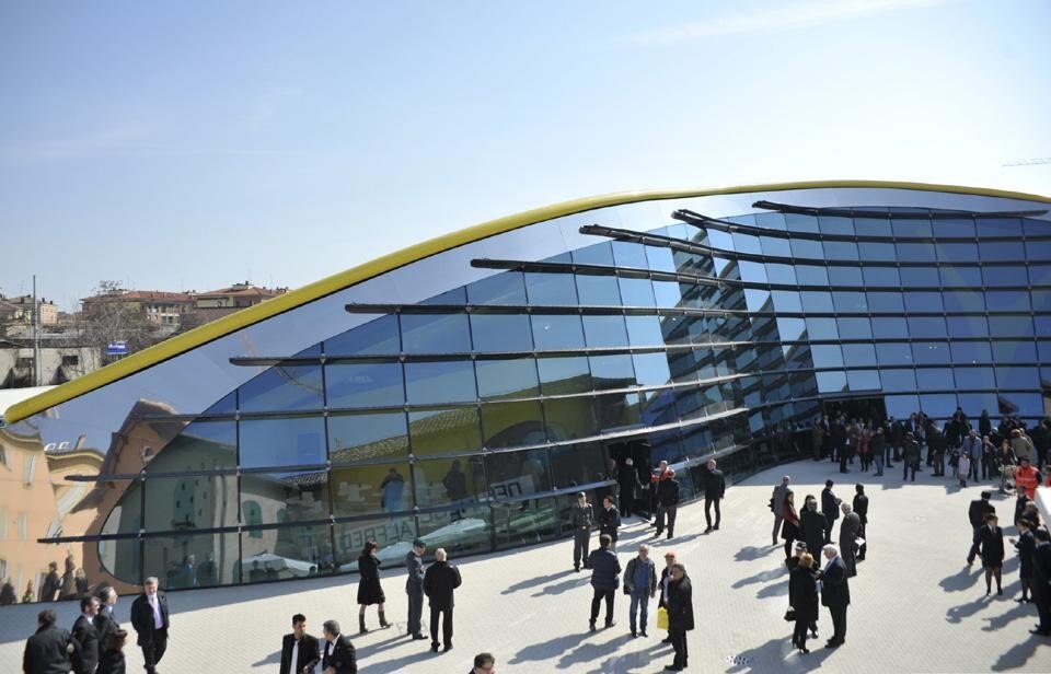 L'ingresso alla Galleria dalla piccola corte che si viene a creare grazie alle curve descritte dalla facciata che abbraccia delicatamente la casa Natale in un gesto di rispetto e simbiosi culturale. La facciata vetrata e’ doppiamente curva in pianta ed e’ inclinata  con un angolo di 12.5 gradi rispetto al terreno. Ogni lastra di vetro-camera e’ sorretta da cavi pre-tensionati in acciaio capaci ognuno di sopportare 40 tonnellate di tensione.